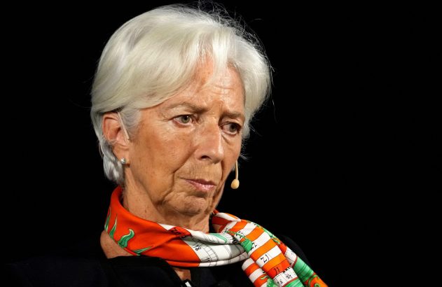 Close-up de mulher de cabelos brancos e lenço colorido, olhando para o lado direito. Há um microfone próximo.