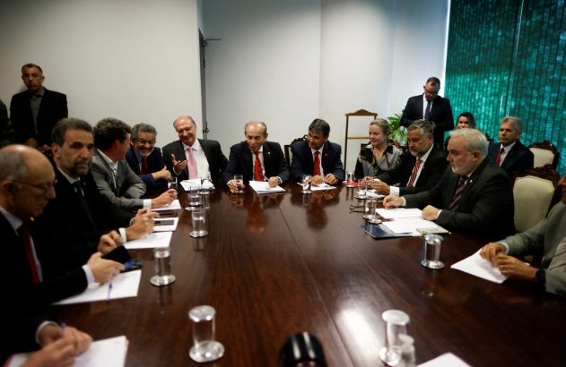 Reunião formal: homens e uma mulher sentados em mesa de conferência com papéis e copos.