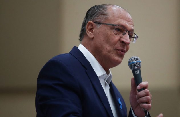 Homem de terno azul e óculos, com broche azul na lapela, fala ao microfone.