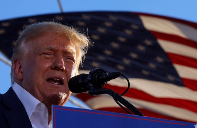 Donald Trump discursando ao microfone com a bandeira dos EUA ao fundo.