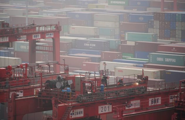 Guindastes vermelhos e pilhas de contêineres coloridos em porto. Trabalhadores soldam em um guindaste, sob neblina.