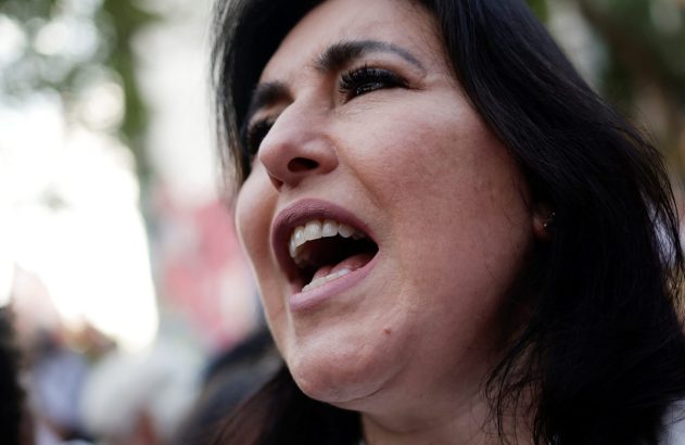 Close-up do rosto de mulher com cabelo escuro, boca aberta e olhar para cima, em fala ou canto.