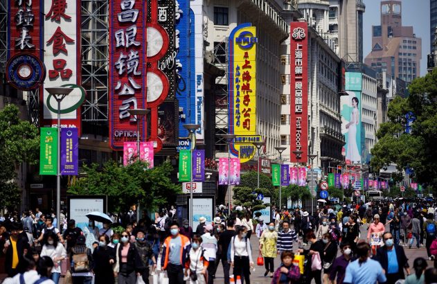 Rua comercial movimentada em Shanghai com pessoas usando máscaras, edifícios altos e letreiros chineses coloridos.