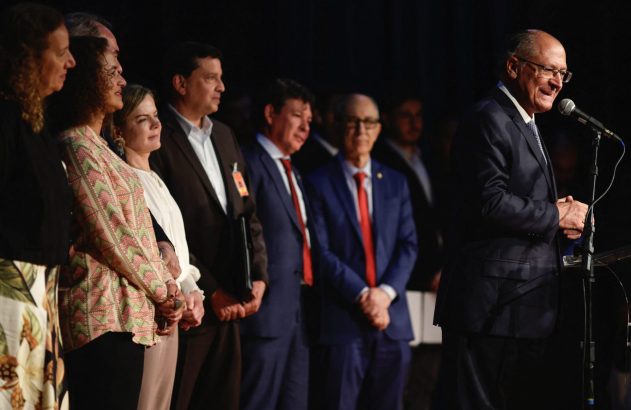 Homem sorrindo discursa ao microfone. À esquerda, grupo de pessoas, incluindo mulheres, ouve atentamente.