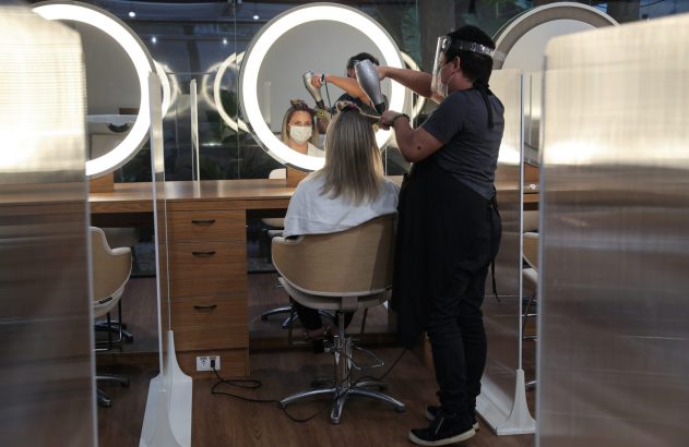 Cabeleireira com protetor facial seca cabelo de cliente em salão. Cliente de máscara no espelho. Barreiras de acrílico visíveis.