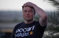 Elon Musk, com camiseta "Occupy Mars", mão na cabeça, em fundo desfocado.
