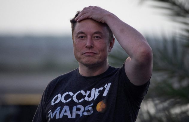 Elon Musk, com camiseta "Occupy Mars", mão na cabeça, em fundo desfocado.