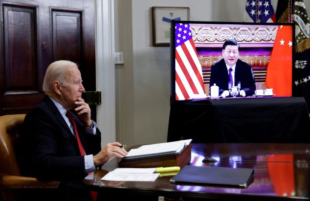 Joe Biden, pensativo, em videochamada com Xi Jinping, que sorri na tela com bandeiras EUA/China.