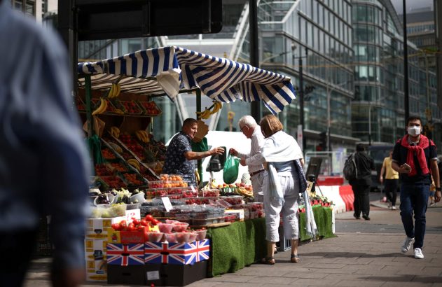 Barraca de frutas com vendedor e clientes. Pessoas caminham, uma de máscara, e prédios modernos ao fundo.