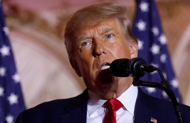 Donald Trump discursa em microfone, com bandeiras dos EUA ao fundo.