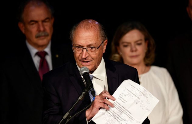 Geraldo Alckmin, de óculos e terno, fala em microfone, segurando papéis, com pessoas ao fundo.