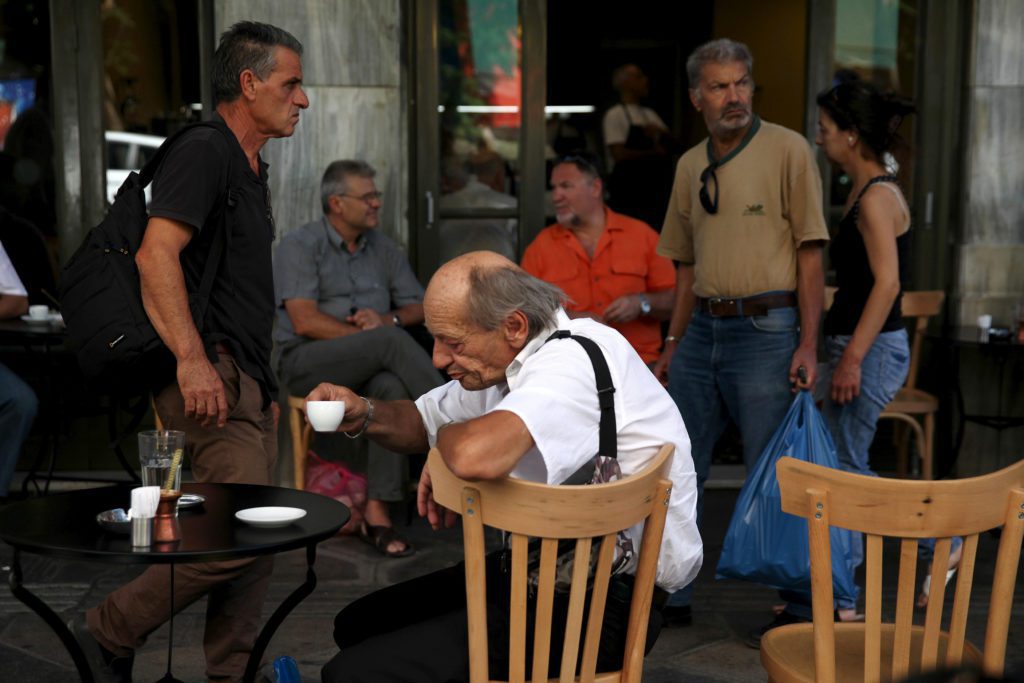 Um idoso segura uma xícara, e um homem passa com mochila, em um café ao ar livre.