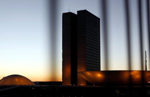 Silhueta do Congresso Nacional do Brasil ao entardecer, com cúpula e concavidade iluminadas, atrás de grades.