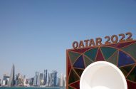 Sinal 'QATAR 2022' com a skyline de Doha e o mar ao fundo, e parte de uma estrutura branca na frente.