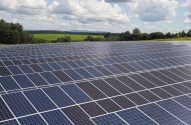 Extensa usina solar com fileiras de painéis. Em campo aberto e paisagem rural, com árvores e céu parcialmente nublado.