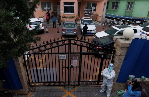 Pessoas em trajes protetores e civis em pátio residencial com carros, portão e placas de restrição.