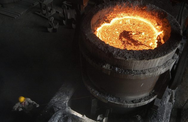 Operário com capacete amarelo manipulando metal incandescente em um grande cadinho na fundição.