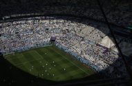 Estádio da Copa 2022: torcida argentina nas arquibancadas, jogadores aquecem no gramado sob luz e sombra.