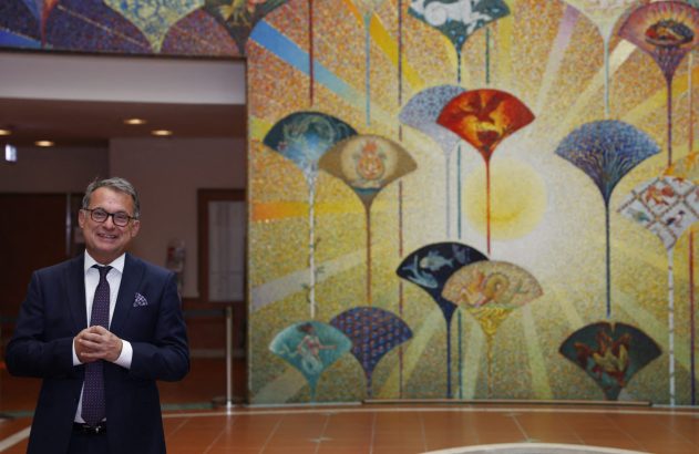 Homem de terno sorrindo, com mural de mosaico colorido de leques e símbolos ao fundo.