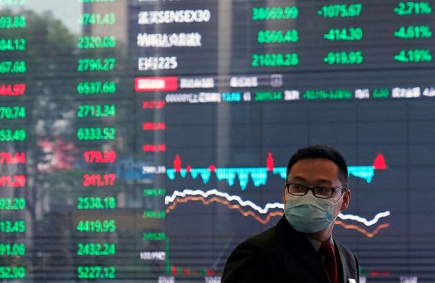 Um homem de máscara está em frente a um grande painel digital exibindo gráficos e números do mercado financeiro.