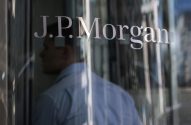 J.P. Morgan refletido em vidro, com uma pessoa de camisa azul em segundo plano.