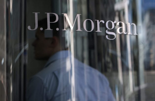 J.P. Morgan refletido em vidro, com uma pessoa de camisa azul em segundo plano.