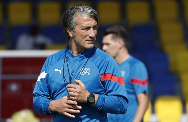 Homem com camisa azul e cabelo grisalho segura cronômetro, olhando para a direita. Fundo de estádio.