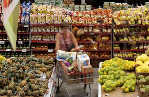 Pessoa empurrando carrinho cheio de compras em atacado. Prateleiras com pães e frutas como abacaxi e limão.