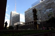 Dois prédios altos sob céu azul, um com antena. Letreiro vertical "Sesc", bandeira do Brasil e pessoas em praça movimentada.
