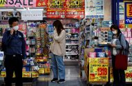 Pessoas usando máscaras em rua comercial no Japão, com um homem ao telefone e duas mulheres olhando produtos.