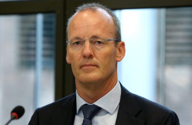 Homem de óculos, terno escuro e gravata, olhando diretamente. Microfone à esquerda sugere evento ou apresentação.