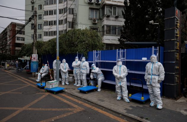Um grupo de pessoas com trajes de proteção brancos em uma rua urbana com estantes azuis, edifícios e carrinhos.