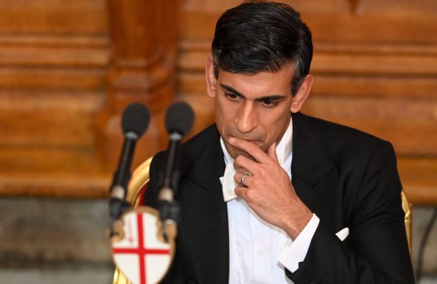 Um homem de smoking e gravata borboleta branca, com a mão no queixo, parece pensativo em frente a microfones.