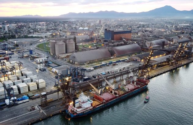 Porto de carga ao crepúsculo: navio atracado, contêineres, silos. Cidade e montanhas ao fundo.