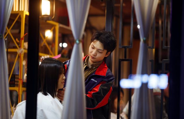 Cabeleireiro com jaqueta colorida corta cabelo de mulher em salão, visto através de cortinas brancas.