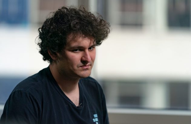 Homem com cabelo escuro e encaracolado, camiseta preta, expressão séria, em ambiente interno desfocado.