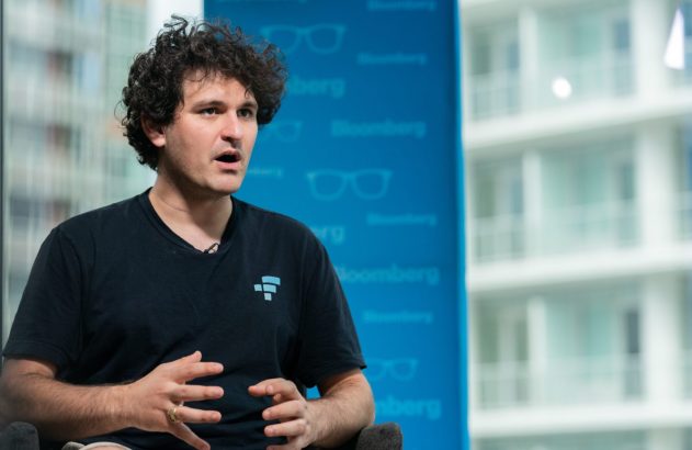 Homem de cabelos cacheados e camiseta preta com logo FTX gesticula enquanto fala em ambiente da Bloomberg.