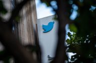 Logo azul do Twitter em um prédio, com folhas e galhos escuros desfocados em primeiro plano.