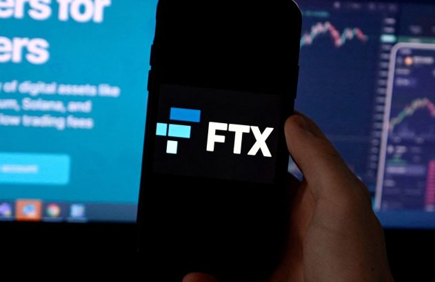 Mão segura smartphone exibindo o logo da FTX. Ao fundo, telas de computador mostram gráficos e texto sobre criptomoedas.