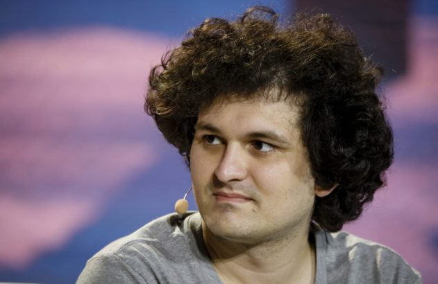 Homem com cabelo cacheado em camiseta cinza e microfone, olhando para o lado. Fundo desfocado rosa e azul.