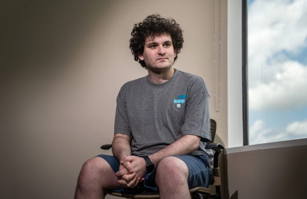 Homem de cabelo cacheado, camiseta cinza e bermuda azul, sentado em cadeira, olhando para o lado. Janela com nuvens ao fundo.