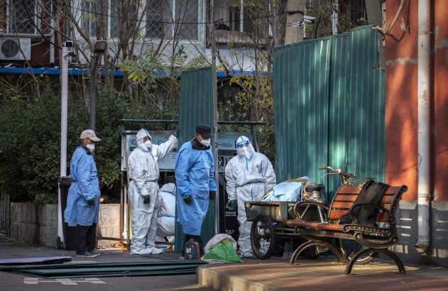 Grupo de pessoas em trajes de proteção numa rua, junto a uma barreira verde, um banco e carrinho.