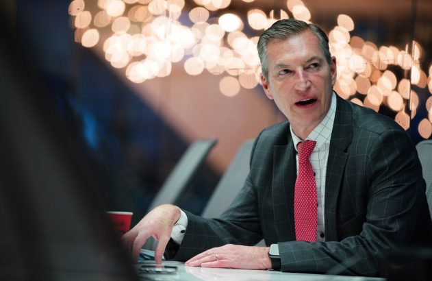 Homem de terno cinza e gravata vermelha, sentado, falando em ambiente iluminado com bokeh.