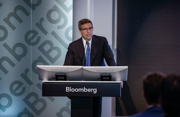 Homem de terno e óculos discursa em púlpito Bloomberg com monitores. Fundo cinza com a marca repetida.