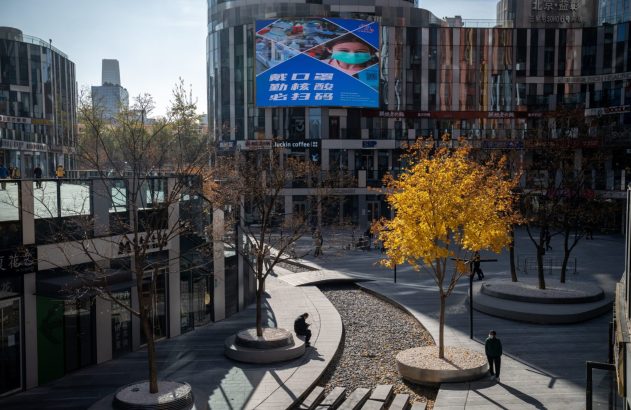 Praça urbana em Pequim com prédios modernos e billboard mostrando criança de máscara. Árvores amarelas de outono e poucas pessoas.