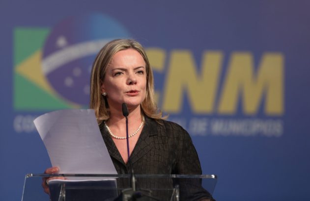 Mulher em púlpito discursando ao microfone, segurando papéis. Fundo azul borrado com logo do Brasil.