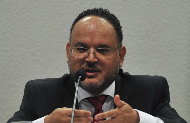 Homem de óculos, terno e barba fala ao microfone, gesticulando com as mãos.