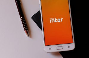Smartphone branco exibe app "banco inter" em tela laranja, com caneta escura ao lado em superfície branca e preta.