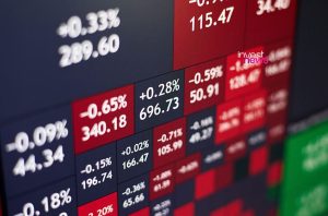 Monitor exibe cotações de mercado com números e percentagens em caixas vermelhas (quedas) e escuras (altas).