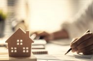 A imagem mostra uma casinha de madeira em primeiro plano, a mão de uma pessoa segurando uma caneta em cima de papéis, exemplificando bem cálculos para avaliar um investimento imobiliário. (Foto: Adobe Stock / Godshutter)